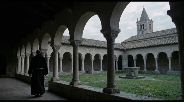 A small cloister at dawn; a monk in black walks away along the arcade.