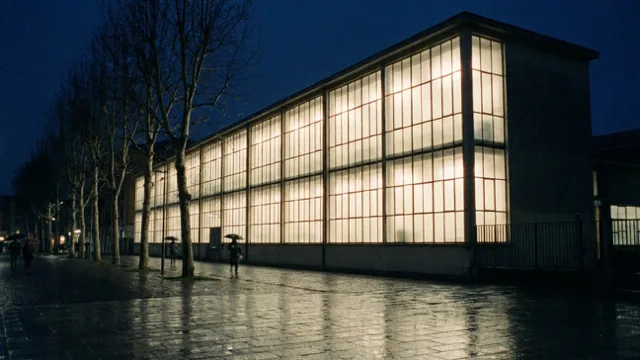 A long modernist glass factory front at night, lit windows doubled in wet paving.