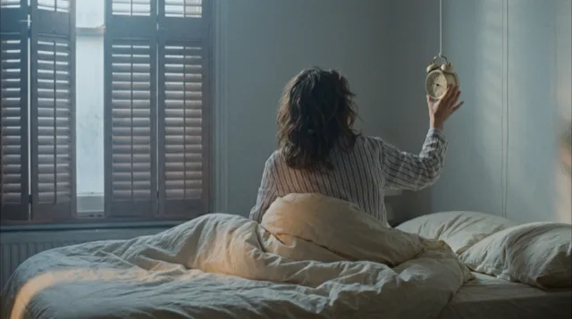 From the foot of the bed: an arm stretches up from the duvet toward the clock, now out of reach.