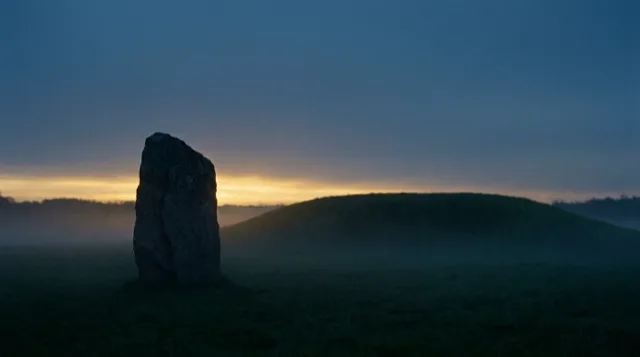 The standing stone between the mounds at dusk, a silhouette against the last gold.