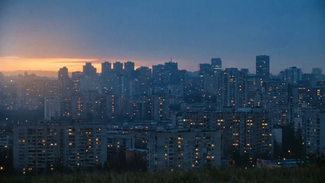 A city at dusk from high above; windows light up one by one.