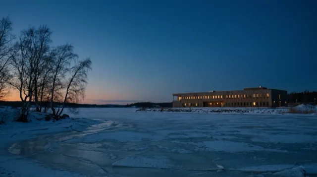 Snow drifts across a frozen Nordic bay at dusk; a low building's windows glow on the far shore.