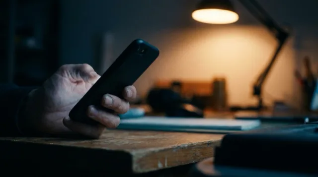 A hand holds a phone at the desk's edge, screen away, its glow on the fingers.