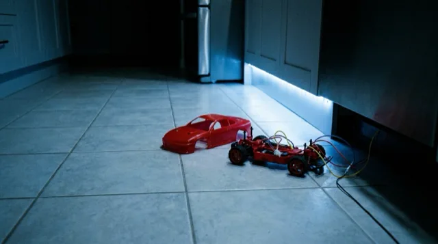 A kitchen floor at night: a red toy car with its shell off, at floor level.